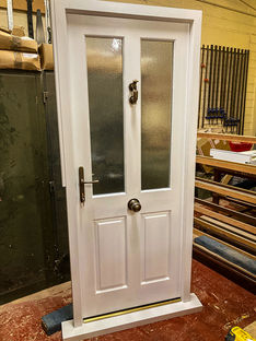 Another timber door, nearly identical in design to the previous, stands in a different workshop location. It features two frosted glass panels and two raised lower panels, with classic door furniture including a round doorknob, letterbox, and traditional knocker. The surrounding workshop shows stacked wood, clamps, and equipment, providing a glimpse into the bespoke joinery environment where the door was constructed.