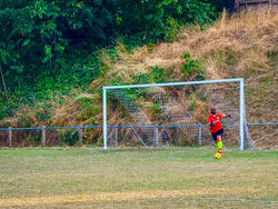 Goalkeeper in a red jersey and neon yellow socks prepares to kick a yellow football from the goal area, with a backdrop of dry grass bank and dense green trees.