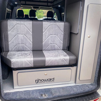 Rear interior view of a camper van showing a compact bench seat with “rjhoward bespoke conversions” branding. The layout includes overhead storage, clean wall panels, and rear access to the main living space.