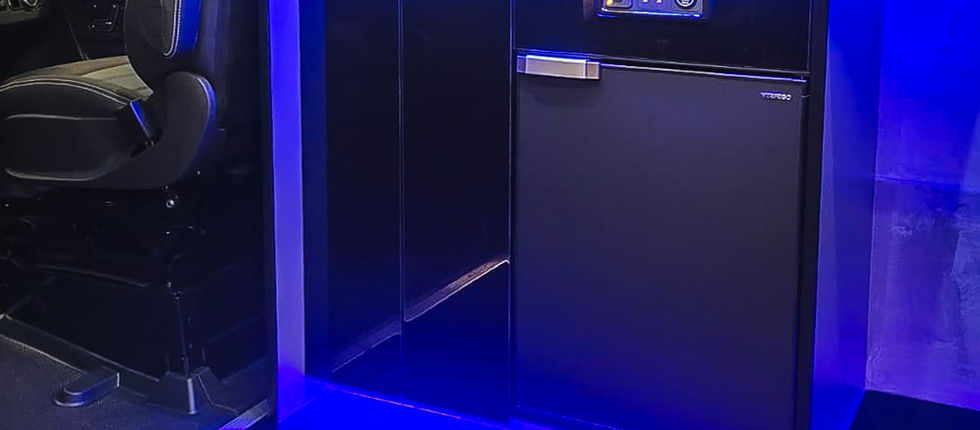 Compact kitchen and storage area positioned immediately behind the cab, featuring integrated refrigeration, practical cupboards, a dark worktop and convenient electrical and USB charging points. A fitted window blind provides privacy when required, while blue LED lighting beneath the cabinetry creates a striking finish and visually connects the kitchen with the rest of the bespoke SC Sporthomes interior.