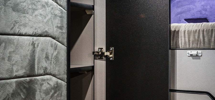 A close-up of a storage cabinet inside the camper van reveals premium materials and craftsmanship. The cabinet features a black door with silver hinges, set against soft gray, padded walls. Inside, neatly organized shelves provide storage for essentials. A small speaker and LED lighting are visible in the background, near an elevated sleeping area with a soft, gray covering. The design blends comfort and practicality, with every space utilized for efficient mobile living.
