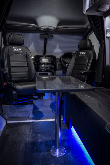 A view from the sleeping loft in a camper van, looking down onto the main cabin area. The image reveals a spacious setup with a large leather seating area, a wall-mounted screen, and a compact microwave. The design includes dark, polished surfaces and ambient blue lighting, creating a stylish and high-tech ambiance. This perspective emphasizes the van’s multi-level functionality, showcasing a smart use of space in a luxurious mobile environment.
