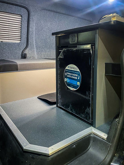 Interior view of a compact camper van featuring a raised speckled vinyl floor with metal trim and a built-in kitchenette unit. A black Vitifrigo fridge is integrated into the unit, with a sticker promoting its cooling efficiency. Next to it is a grey cushioned bench with a shuttered side window above. The walls are lined in grey carpet material, and LED lights on the ceiling illuminate the space, highlighting the neat, functional layout.