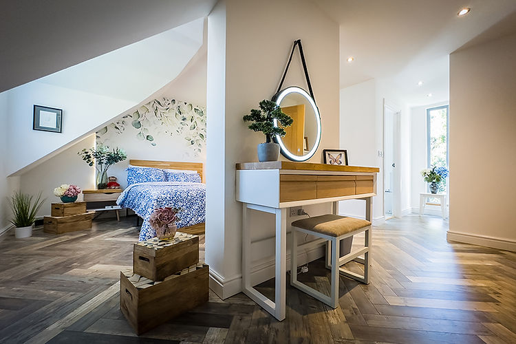 The image shows a modern, well-lit bedroom and dressing area with a cozy, inviting ambiance. To the left, a bed with a blue floral duvet is positioned against a wall adorned with a leafy mural. The angled ceiling above adds a unique architectural touch. On the right side of the room, a white vanity with a round, backlit mirror and a cushioned stool sits near a door leading to a bright corridor. Decorative touches include plants, wooden crates used for storage, and floral arrangements in vases. Light wood flooring in a herringbone pattern extends throughout, adding warmth to the overall clean, contemporary design.