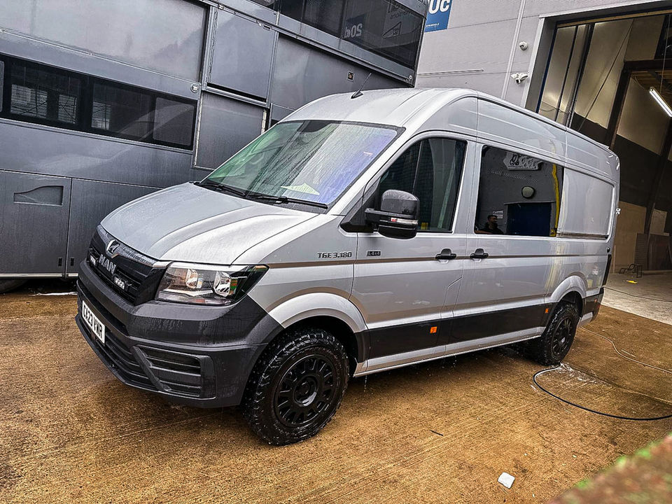 Exterior view of a silver MAN TGE 3.180 4x4 van, fully converted by RJ Howard Bespoke Conversions. The van is parked outside a workshop on wet concrete, featuring all-terrain tires, black alloy wheels, and a high roof ideal for camper applications. Branding and 4x4 badging on the side reflect the van’s rugged capability and suitability for off-road or overland travel. Tinted windows and a sleek two-tone paint finish reinforce a professional and adventure-ready appearance.