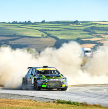 The RMG Groundworks Citroën rally car powers through a dusty corner during a high-speed stage, creating an enormous cloud of dust across the circuit behind it. The vivid green, black and purple car remains sharply visible at the centre of the action as it changes direction. Open fields and distant farmland provide the backdrop to a dramatic image filled with speed, movement and rallying intensity.