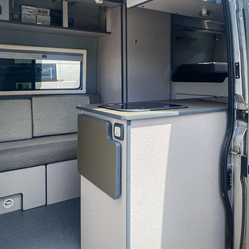 Side entry view into a camper van, showing a small kitchen counter with black-glass hob and soft-close cabinets. Opposite is a minimalist bench seat under a window, with open shelving above containing electrical control panels.