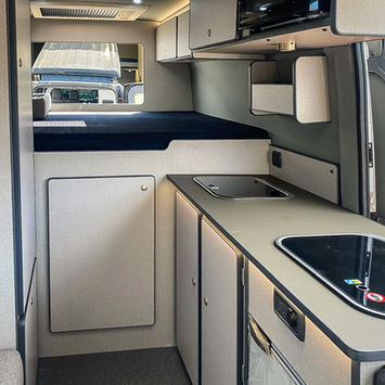 Rearward view of the camper van’s interior from the kitchen, showing a raised bed platform, overhead cabinets, and soft lighting. The space is optimized for function and comfort, with a cohesive color palette of light grey and matte black.