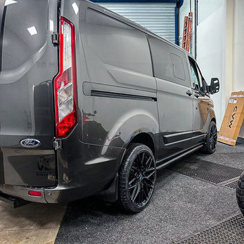 Rear three-quarter view of a dark gray Ford Transit Custom panel van inside a workshop. The vehicle is fitted with black alloy wheels, side steps, and a subtle rear spoiler above the rear doors. Its smooth body panels, privacy glass, and metallic paintwork create a clean, modern look, while the tall rear taillight and long-wheelbase proportions emphasize its practicality and cargo-carrying capability.