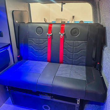 Rear passenger bench seat inside a bespoke camper van conversion, upholstered in black leather with patterned inserts and bright red seatbelts. Blue ambient lighting highlights the surrounding cabinetry and interior trim, creating a premium and contemporary atmosphere.