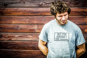 A picture of Guy Martin in front of a wooden plank background, smiling downward with a playful expression. He wears a light grey T-shirt with the humorous text "NOT FOR AIRCRAFT USE.SIDEBURN" in a bold font across the chest. His casual stance, coupled with curly hair and a natural, relaxed demeanor, conveys a sense of approachability and lightheartedness. The backdrop's rich wood texture adds warmth to the image, complementing the friendly vibe of the scene.