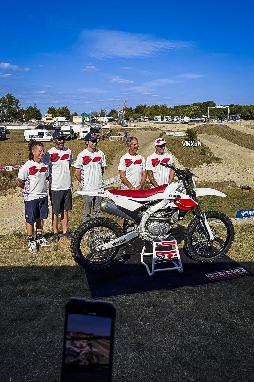 Five men stand proudly beside a red and white Yamaha motocross bike on a stand. They wear matching white shirts with bold red logos. The dirt track and spectator area stretch out behind them under a bright blue sky, with “VMXdN” signage in the background.
