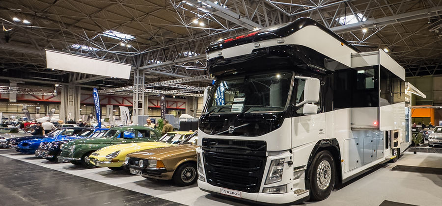 Wide shot of an exhibit hall featuring a massive black-and-white Volvo motorhome parked alongside a row of classic cars. The motorhome’s towering presence and sleek design contrast with the vibrant colors of the vintage vehicles, creating a striking juxtaposition between classic and modern styles.