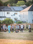 Close-up of the black-and-white striped team huddled near their dugout, with substitutes sitting and others drinking water. A goalkeeper in a bright blue kit stands among them, while the setting sun casts a warm glow on nearby houses.