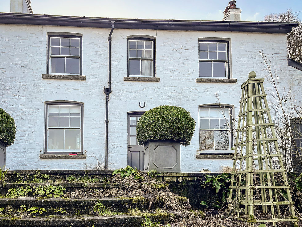 Close-up of the cottage’s front elevation, showing whitewashed stone walls, sash windows with grey trim, and a modest front door. The stone steps leading up to the entrance are edged with moss and overgrown greenery. Topiary shrubs and a wooden garden obelisk add structure to the natural charm.