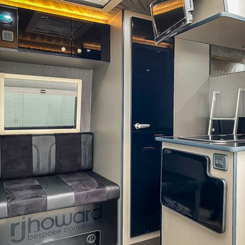 Interior view of a luxury camper van showing a black and grey upholstered bench seat beneath a window with a pleated blind. Above are glossy black overhead cabinets with a digital control panel and thermostat display. To the right, a compact kitchen unit with a sink and flip-up worktop extension sits beside a small metal ladder leaning against the wall. A flat-screen TV is mounted above the kitchen near the ceiling.