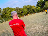 Coach in a red “MARDY FC” shirt stands on the sideline with arms folded, watching the match unfold across the pitch under a summer sky.