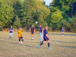 Midfield action as a player in a yellow jersey and black shorts tracks a high ball, while navy-and-light-blue opponents and a referee watch under bright sunlight.
