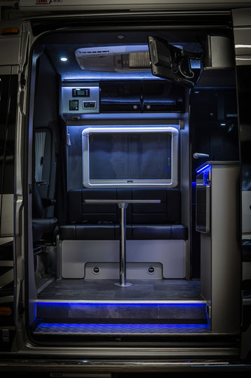 View inside a modern camper van featuring a sleek black-and-silver interior. The space includes a compact dining area with a cushioned bench, mounted table, overhead control panels, and a window with a built-in shade. Cool-toned LED lighting highlights the ceiling and underfloor edges, giving the space a high-tech and inviting ambiance.