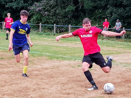 A football player in a red Mardy FC jersey and black shorts swings his leg to kick a ball on a dry, patchy grass pitch, while an opposing player in a blue and yellow kit runs behind him. Several teammates and spectators are visible in the background near the touchline and trees.