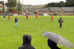 Spectators in rain jackets and umbrellas watch from the touchline as players in red and navy prepare for play on a wet village football pitch.