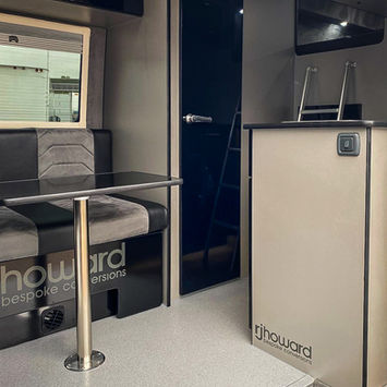 Interior view of a modern camper van showing a compact lounge and kitchen area. A two-tone bench with “rjhoward bespoke conversions” branding sits beside a black table with a metal pedestal. A glossy black door leads to the rear, and a ladder is mounted next to a kitchen counter with integrated appliances and overhead storage.