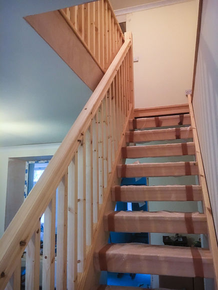 Upward view of a newly installed timber staircase with vertical pine balusters and a handrail on both sides. The treads are covered in protective plastic and brown tape, and the stairs lead to a landing with matching balustrade.