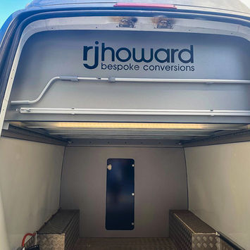 Interior rear view of a converted camper van featuring a clean cargo area with diamond-plate metal benches on both sides. Above, the upper wall displays the “rjhoward bespoke conversions” logo, along with fitted rails and overhead lighting. The walls are light-colored, and the space is uncluttered.