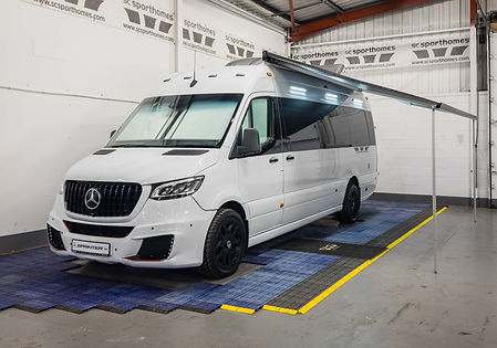 A full front-and-side view of the Mercedes-Benz SCS Sprinter 2025 with its awning fully extended. The sleek van is parked on a rubberized mat inside the “SC Sporthomes” showroom. The black grille with the Mercedes emblem, large tinted windows, and high-quality black alloy wheels enhance its premium aesthetic. The extended awning showcases its suitability for outdoor and recreational purposes.
