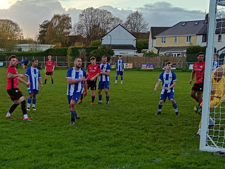 A dynamic football match unfolds on a grass pitch as players in red and blue kits react to a moment near the goal. Several players in blue-and-white striped jerseys watch the ball intently, while red-shirted opponents move into position, creating a sense of anticipation and tension. The goalkeeper, dressed in yellow, is partially obscured as he attempts to defend the near post. Behind the action, houses, trees, and a glowing sunset sky frame the scene, adding depth and atmosphere. The mixture of focused faces, quick movements, and varied body language captures the fast, unpredictable nature of grassroots football.