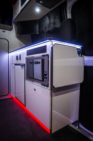 A minimalist kitchenette featuring a compact oven, refrigerator, and sleek cabinetry. LED lighting runs along the countertop edges and base, adding a futuristic ambiance to the space.
