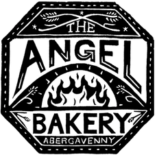 Vintage-inspired black-and-white logo for The Angel Bakery Abergavenny, framed within an ornate hexagonal border. Decorative wheat motifs and a stylized oven flame at the center evoke traditional baking, artisan craftsmanship, and locally made bread.