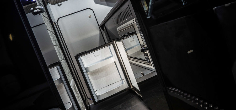 The kitchenette inside the camper van features a sleek, modern design. The black cabinetry houses an open refrigerator with white shelving and a compact oven with a stainless steel door. The reflective surfaces of the cabinets and appliances contrast with the soft gray, padded walls. A small sleeping area is visible above the kitchenette, softly illuminated with subtle LED lighting, showcasing the van's efficient use of space for cooking and storage.