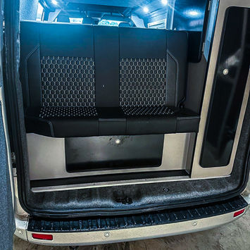Rear view inside a camper van showing a two-seat bench with dark hexagonal-patterned cushions and overhead lighting. The back doors are open, revealing the textured interior trim.
