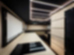 Wide-angle interior shot of a high-end camper van showing a minimalist kitchen with an induction cooktop, smooth beige countertops, and a modern black ceiling with geometric LED lighting. The view extends toward a rear lounge with cushioned seating.