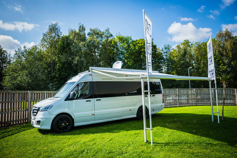 An exterior view of the camper van with an extended awning creating a shaded outdoor area. The sleek white vehicle is parked on green grass, framed by trees and fencing, with branded flags completing the setup.