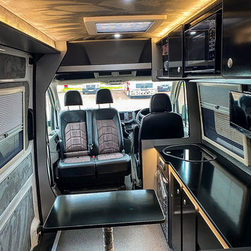 Perspective from the rear living area of a camper van conversion facing the cab. The interior showcases a central table with a wrapped leg, swivel front seats with matching grey-and-black upholstery, and a compact kitchenette on the right featuring a sink, fridge, and ample storage. The ceiling has ambient LED lighting and a skylight. Modern, streamlined design with a blend of functionality and comfort is evident throughout.