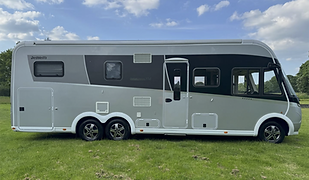 A sleek, modern Dethleffs Globebus XL I motorhome is parked on a lush green field under a partly cloudy blue sky. The motorhome has a stylish two-tone exterior in silver and dark grey, with smooth, aerodynamic curves and multiple windows along its side. The design features clean lines, twin rear axles for added stability, and several exterior access panels and vents, indicating various built-in utilities. The front cab area includes large wraparound windows, suggesting a panoramic driver’s view. The open grassy area and tranquil setting emphasize freedom and outdoor adventure, making this vehicle ideal for luxurious off-grid travel or extended road trips in comfort.