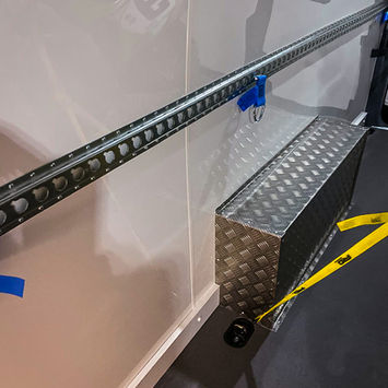 Close-up of a diamond-plate wheel arch box and grey non-slip flooring, with a yellow tie-down strap attached to the integrated floor ring. A Kawasaki bike number plate is partially visible in the reflection on the gloss wall.