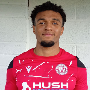 A football player poses for a portrait in front of a white brick wall, wearing a red team jersey with black shoulder accents. The shirt displays the logo of HUSH Soundproofing and a circular club crest reading “Mardy Football Club.” The player stands confidently, looking directly at the camera with a slight smile, showing a calm and composed expression. The image appears to be an official team photo, highlighting the player’s uniform and club affiliation.