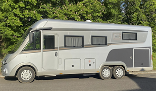 A large, luxury motorhome is parked on a sunlit road with a backdrop of leafy green trees. The vehicle is a Carthago Chic E-Line, finished in sleek silver and grey with subtle bronze and charcoal accents along the sides. It features an aerodynamic front with panoramic windows, flush-mounted doors, and tinted rectangular windows with interior curtains drawn. The body includes multiple external access compartments and utility panels, with twin rear axles for improved stability and support. The clean lines, upscale finish, and thoughtful design suggest a high-end touring experience, ideal for extended travel in comfort and style, blending seamlessly into both natural and urban settings.