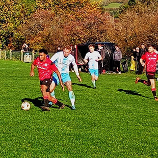 In the middle of an amateur football match, a player in a red and black kit drives the ball forward while being closely pursued by a defender in a light blue and white kit. The two athletes are in motion on a grassy pitch, with additional players, team staff, and spectators visible in the background. A black equipment tent and trees with colorful autumn leaves line the edge of the field. The image conveys a sense of urgency and competition as players chase possession in bright daylight, framed by the vibrant natural surroundings of a community sports ground.