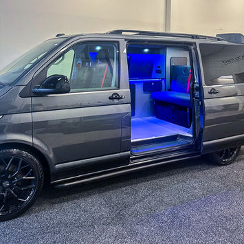 Side view of a Volkswagen camper van with the sliding door open, revealing a custom conversion interior illuminated by blue ambient LED lighting. Inside, a bench seat, integrated cabinetry, storage compartments, and camper equipment are visible, creating a modern and inviting living space. The contrast between the sleek gray exterior and vibrant blue interior lighting emphasizes the premium craftsmanship of the conversion.