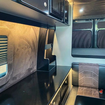 Close-up view of the camper van kitchen and living area, showing glossy black cabinets, a microwave, and floating shelves above a counter workspace. Soft suede-style wall coverings and ambient lighting enhance the luxurious yet compact design. The mauve and black bench seat sits adjacent to the cabinetry, forming a comfortable, cohesive space.