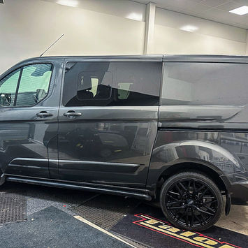Full side profile of a dark gray Ford Transit Custom van displayed inside a workshop. The van features tinted side windows, black multi-spoke alloy wheels, side steps, and a sleek metallic finish that reflects the surrounding lighting. A compact side window is integrated into the sliding door area, while the clean body lines and lowered stance contribute to a refined, contemporary appearance. The combination of practical van proportions and custom styling elements creates a versatile vehicle suited for commercial use, camper conversion, or everyday transport.