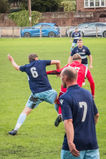 A player in a navy kit lunges into a strong tackle against an opponent in red, while teammates look on during an intense midfield battle.