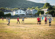Players from both teams compete for an aerial ball, with one in a black-and-white striped kit jumping high and arms raised. Red-shirted opponents challenge nearby, while the referee watches closely in the center of the pitch. A cloudy sky and leafy trees frame the scene.