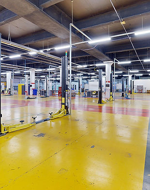 This expansive industrial space is characterized by high ceilings with exposed beams and brightly lit by numerous fluorescent lights. The floor is painted in a vibrant yellow with guiding lines in multiple colors, suggesting designated work or walk areas. Various lift stations with tall, black vertical posts and yellow bases are evenly distributed throughout the space. The area is largely open with a few pieces of machinery and workbenches around the periphery, indicative of a setting prepared for technical or mechanical work. The environment is clean and orderly, ready for productive activity. The presence of safety signs and absence of clutter reflect a commitment to organization and safety standards in the workplace.