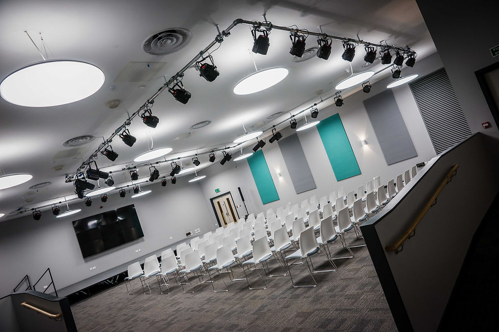 The image shows a spacious and modern auditorium with a descending arrangement of white chairs facing a stage area with a presentation screen. The ceiling is fitted with a combination of round and rectangular lights, and a track of stage lighting systems with multiple spotlights. There are rectangular sound dampening panels on the side walls, alternating with blocks of teal color that add a vibrant contrast to the otherwise neutral color palette of the room. The carpeted flooring is gray with speckled patterns, and there is a stairway with a golden handrail on the right leading down to the stage area. This setting appears to be well-equipped for conferences, lectures, or performances, with its professional lighting and sound systems, and an overall clean and contemporary design.