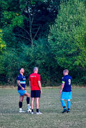 Two footballers in navy kits and a coach in a red “Mardy FC” shirt talking on the field, with one player resting a foot on the ball and a backdrop of leafy trees.